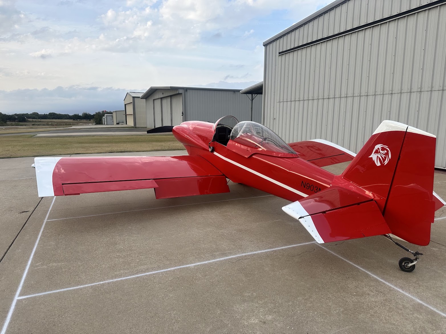 A red and white RV tailwheel aircraft parked on the ramp in front of a hangar